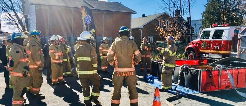 Un instructeur explique une manœuvre d'alimentation en eau à un groupe de pompiers, près d'un bassin portatif et d'une autopompe